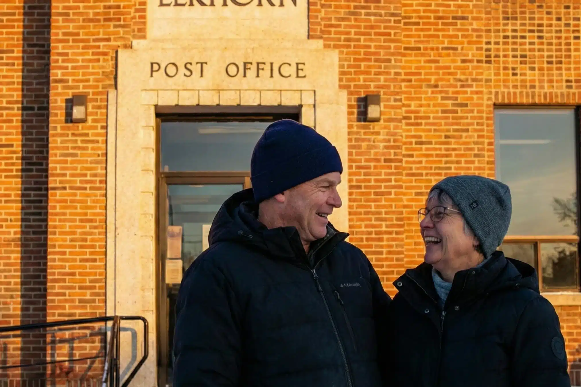 A mother and adult daughter smiling and walking together on a small-town street in Rivers, Manitoba, with local buildings and a parked truck visible in the background.